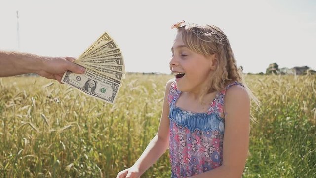 A young girl is given banknotes of dollars and she is very happy about it against a background of nature in a warm sunny day. Money, finances and people concept - smiling little european girl with