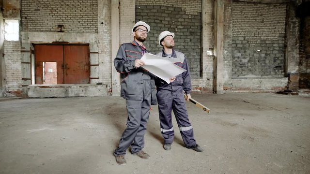 Two men dressed in uniform and safety hardhats are standing on building site and looking over working area. Foreman is holding contsruction plan. Builder is holding spirit level.