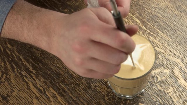 Male hand, latte art pen. Frothy coffee on wooden table.
