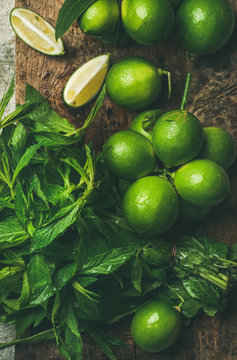 Flatlay of freshly picked organic limes and mint leaves for making cocktail or lemonade on wooden rustic board background, top view, close-up