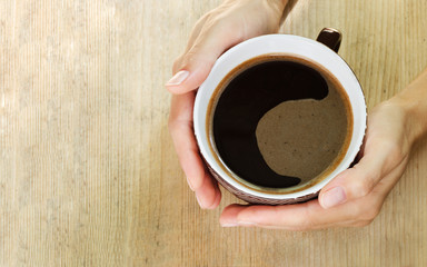 Hands of a woman hugging a large cup of coffee. Wooden background.  Top view. Copy space