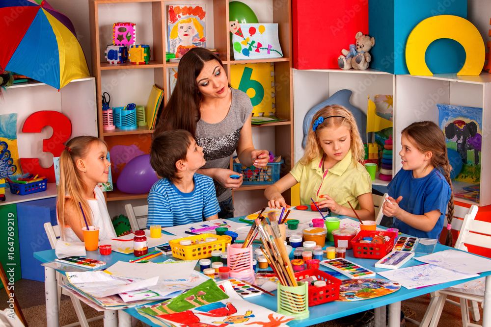 Wall mural Small students with teacher finger painting in art school class. Mom and kids at home. Children with teacher preparing exhibition of children's drawings. Favorite children's teacher.