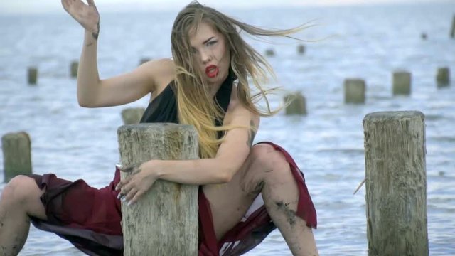 Energetic blonde with long hair in a red dress in the mud beautifully poses in the estuary holding on to wooden poles for salt pools
