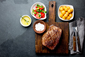 Grilled beef steak served on wooden table with tomato salad and potatoes balls. Barbecue, bbq meat beef tenderloin. Top view, slate background