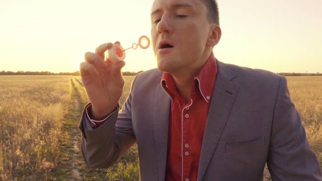 Joyful smiling businessman in a suit on a wheat field  blows soap bubbles and evades them, slow motion