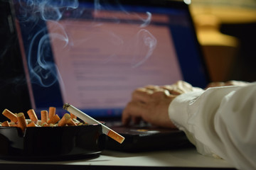 Man working on laptop computer with lit and smoking cigarette on an ashtray filled with cigarette butts