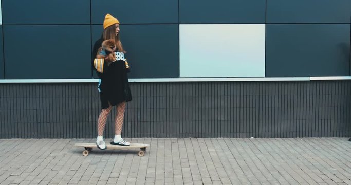 Young stylish Caucasian female riding longboard with her Pomeranian Spitz dog puppy outdoors. 4K UHD RAW edited footage