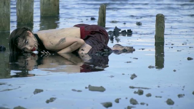 Beautiful blonde smeared in mud in a wet dress, lies in the middle of the estuary, touching her face with water near the wooden posts from the ruined salt pool