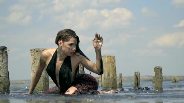 Beautiful blonde smeared in mud in a wet dress, lies in the middle of the estuary near the wooden posts from the ruined salt pool against the blue sky and sharply throws back her hair, slow motion