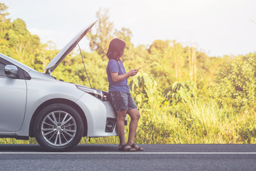 Woman using smartphone in front of her broken car on the road. Contacting car technician or need help concept