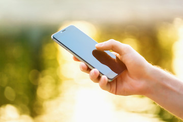 Mockup of male hand holding cellphone with blank screen outdoors in park at golden sunset background.