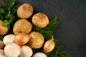 Raw fresh onions, potatoes, garlic, mushroom, greens and seasoning on dark stone background with copy space. Top view ingredients for cooking a delicious cream soup. Healthy vegetarian food.