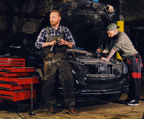 Fototapeta na wymiar Two bearded tattooed mechanics near the car in a workshop.