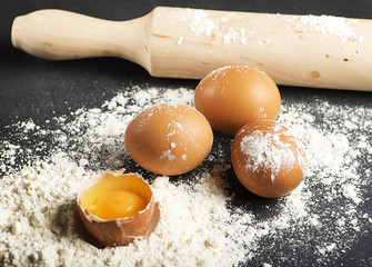 Eggs on flour next to wooden roller on black wooden table. Horizontal studio shot.