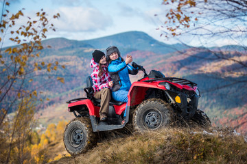 Fototapeta na wymiar Happy women in jackets and hats on red quad bike at the hill makes selfie on the phone with mountains in blurred background. Sunny autumn day