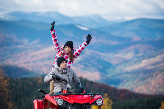 Close-up of man driving atv quad bike, girl sitting behind him and raised her hands up on blurred background of beautiful landscape mountains