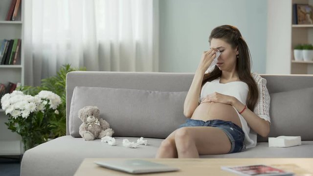Pregnant lady sitting on sofa crying drying tears with tissue, hormonal disorder