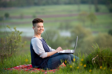 Happy female wearing earphones while working on her laptop sitting on the grass outdoors copyspace technology online connectivity mobility wireless internet project