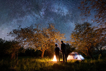 Rear view of romantic couple tourists standing at a campfire, holding hands near tent under trees and beautiful night sky full of stars and milky way. Night camping. Long exprose