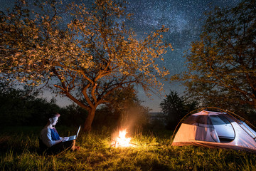 Female tourist using her laptop in the camping at night. Woman sitting near campfire and tent under trees and beautiful night sky full of stars and milky way