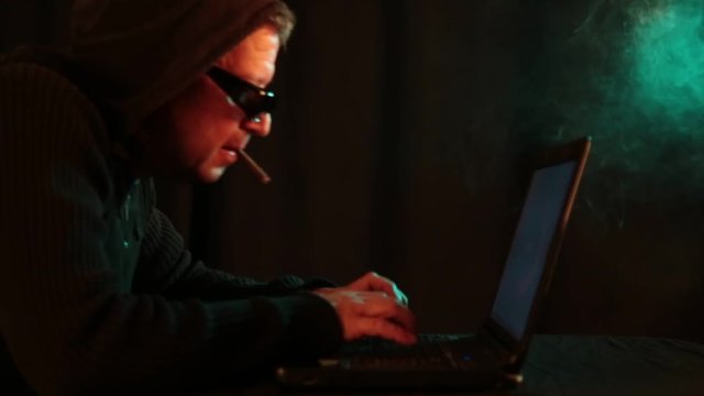 Rauchender Mann mit Sonnenbrille und Hoodie im Homeoffice tippt auf dem Laptop