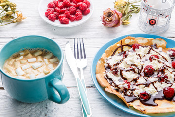 Homemade pancakes or Russian pancakes with chocolate sauce, whipped cream and raspberries on a plate on a white wooden background.