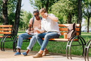 multicultural young students sitting on bench and studying in park together