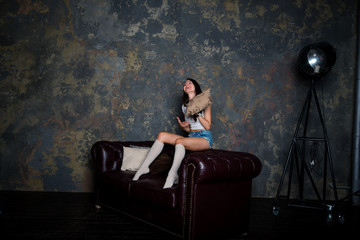 Fototapeta na wymiar Красивая девушка сидит на диване в джинсовых коротких шортах, гольфах и белой майке в интерьерной студии