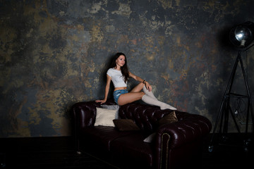 Красивая девушка сидит на диване в джинсовых коротких шортах, гольфах и белой майке в интерьерной студии