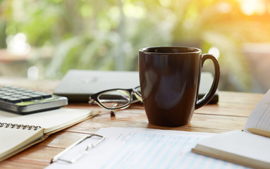 Coffee mug and business objects on wooden desk with greenery background, selective focus