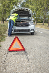 Young man examining his damaged car parked on the side of the road and a red triangle to warn