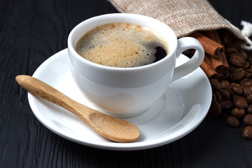 Coffee in a white cup with a saucer and a wooden spoon on a black background with a bag of coffee beans