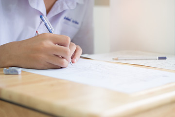 Blurred of Asian boy students hand holding pen writing fill in Exams paper sheet or test paper on wood desk table with student uniform in exam class room, education concept, Thailand