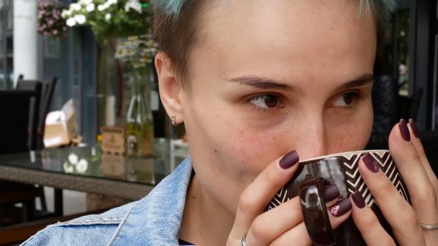 A girl is drinking cappuccino. Video on the veranda of the cafe. A cheerful woman drinks coffee from a beautiful cup. Summer shot.