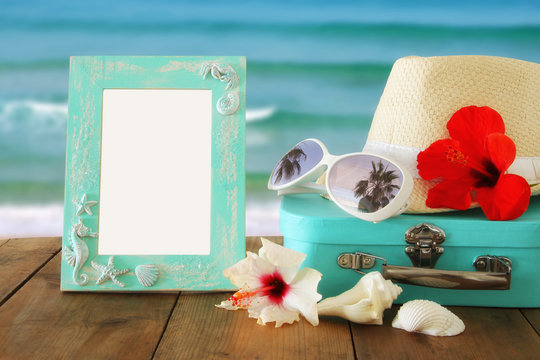 Fedora hat, tropical hibiscus flower next to blank frame over wooden table and beach landscape background