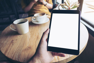 Mock up image of a hand holding black tablet pc with white blank screen on wooden table with woman using mobile phone in background in modern cafe