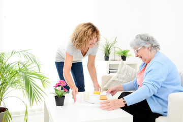 cheerful mature woman serving breakfast and taking care of elderly senior woman at home