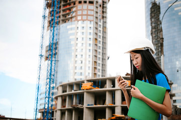 Asian woman in a blue dress and white protective helmet. engineer supervises inspection and control of works at construction site. Business telephone conversations. Communication via smartphone