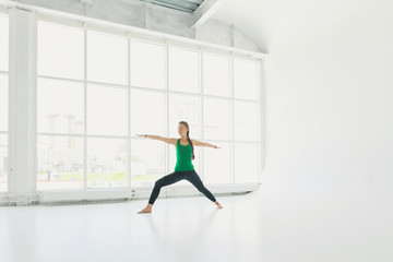 Fototapeta na wymiar Beautiful smiling happy young woman working out indoors, doing yoga exercise near floor window with city view