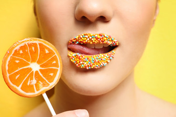 Beautiful young woman holding lollipop near lips covered with sprinkles on color background, closeup