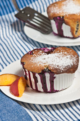 Fresh, baked blueberry-peach cobbler in a on a round, white dish with two slices of peach and a spatula on a blue and white stripped table cloth 