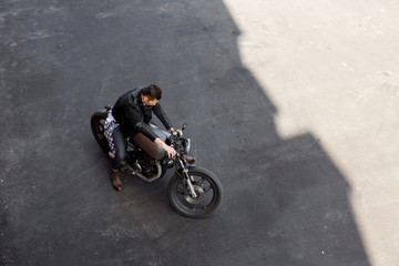 Top view of a handsome rider guy in black biker jacket, checkered shirt, denim and boots ride his classic style cafe racer motorcycle. Bike custom made in vintage garage. Brutal fun urban lifestyle. - 164868615