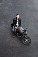 Top view of a handsome rider man in black biker jacket, checkered shirt, denim and boots sit on classic style cafe racer motorcycle. Bike custom made in vintage garage. Brutal fun urban lifestyle.