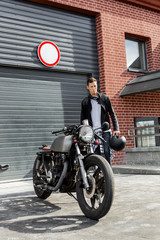 Handsome young rider man in black leather biker jacket go to his classic style cafe racer motorcycle industrial gates as background. Bike custom made in vintage garage. Brutal fun urban lifestyle.