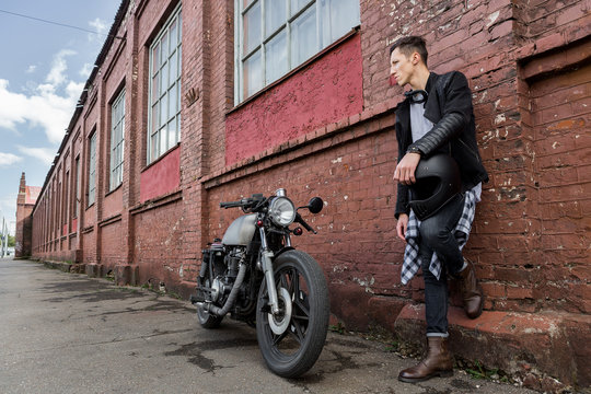 Closeup of a handsome rider biker man hand with black helmet near brick wall and classic style cafe racer motorcycle. Bike custom made in vintage garage. Brutal fun urban lifestyle. Outdoor portrait.