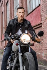 Handsome rider biker man in black leather jacket sit on classic style cafe racer motorcycle and look forward. Bike custom made in vintage garage. Brutal fun urban lifestyle. Outdoor portrait.
