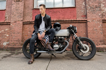 Fototapeta na wymiar Handsome rider biker man in black leather jacket, jeans and boots sit on classic style cafe racer motorcycle. Bike custom made in vintage garage. Brutal fun urban lifestyle. Outdoor portrait.
