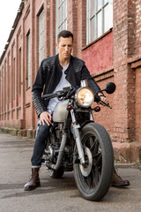 Fototapeta na wymiar Handsome rider biker guy in leather jacket sit on classic style cafe racer motorcycle and check fuel before ride. Bike custom made in vintage garage. Brutal fun urban lifestyle. Outdoor portrait.