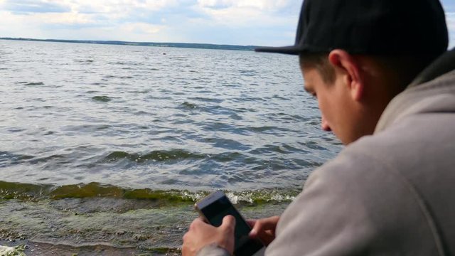 The guy uses the phone while sitting on a chair by the lake. The traveler dials a message on the smartphone.