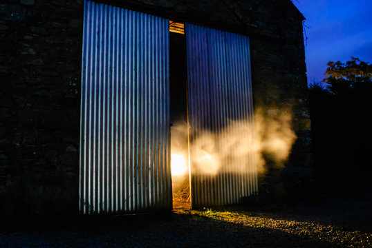 Light shines through smoke coming out from half open doors of an old Irish stone barn at night
